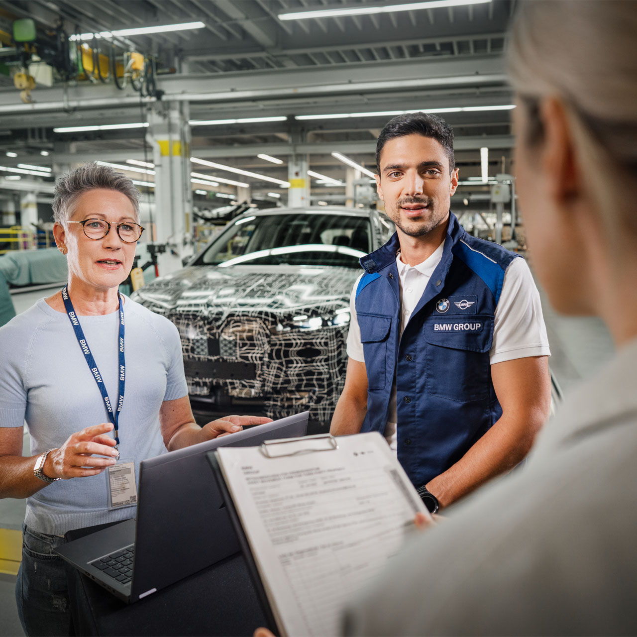 Two people in work attire stand in a BMW factory. One person holds a laptop and the other holds a document. A partially covered vehicle is in the background.