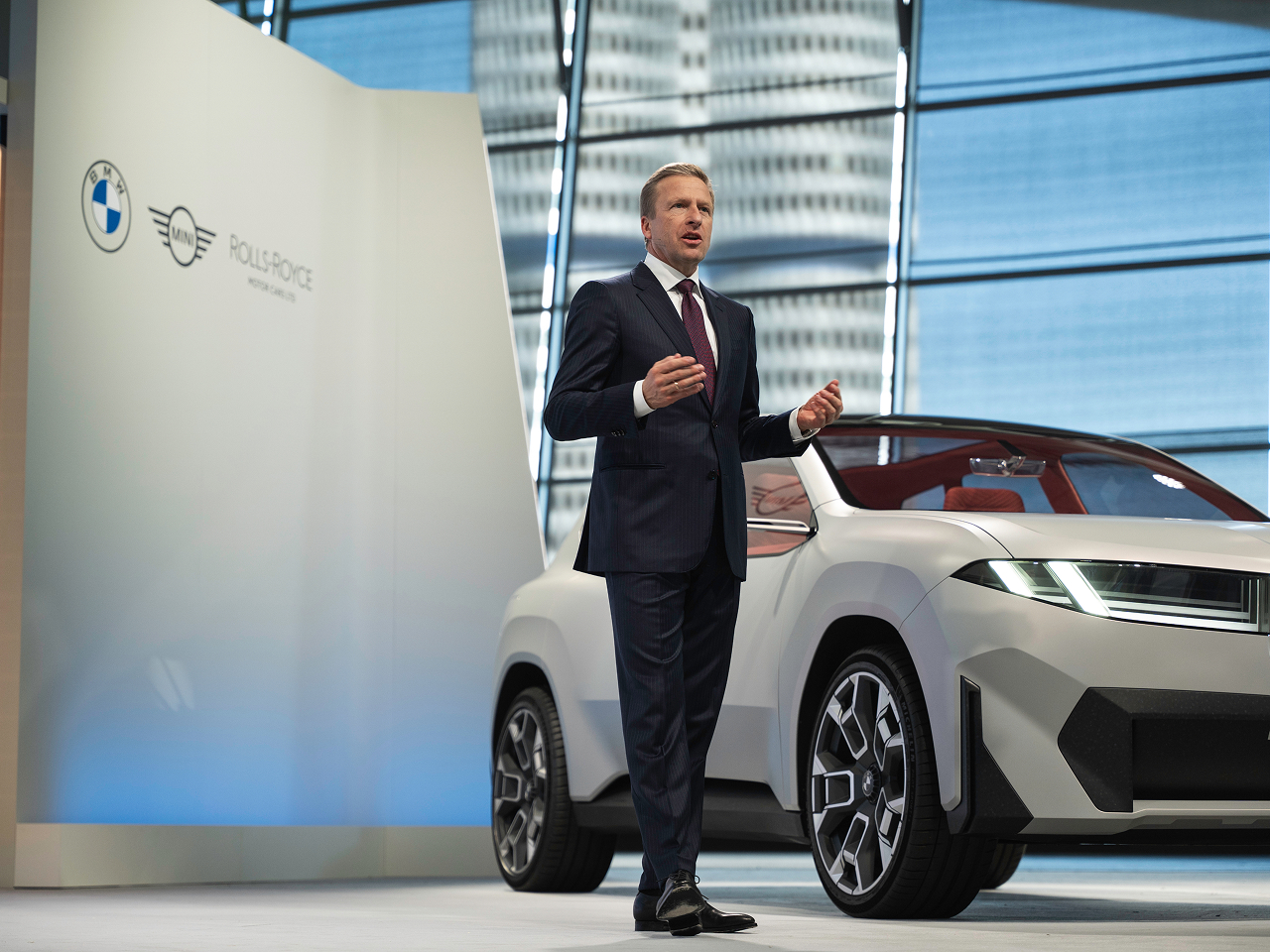 Oliver Zipse speaking on stage next to a silver BMW concept vehicle, with the BMW Four-Cylinder Tower in the background.