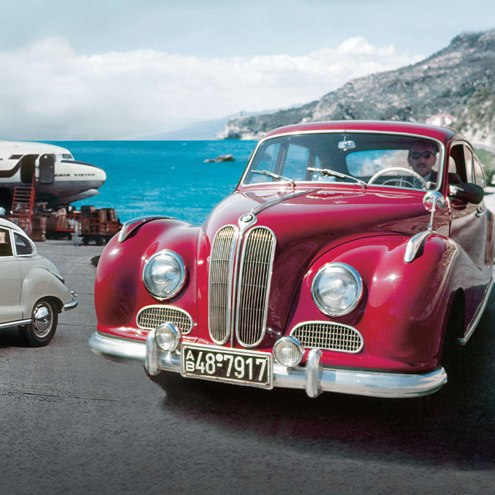 Two BMW 501, the first model produced after the war, one in grey and one in red. In the background there is a plane and the sea.