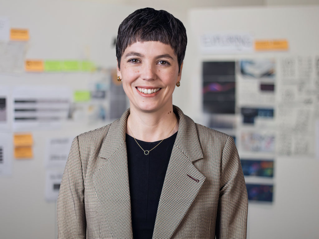 Woman with short dark hair wearing a beige checkered blazer. Various documents and graphics are visible in the background.