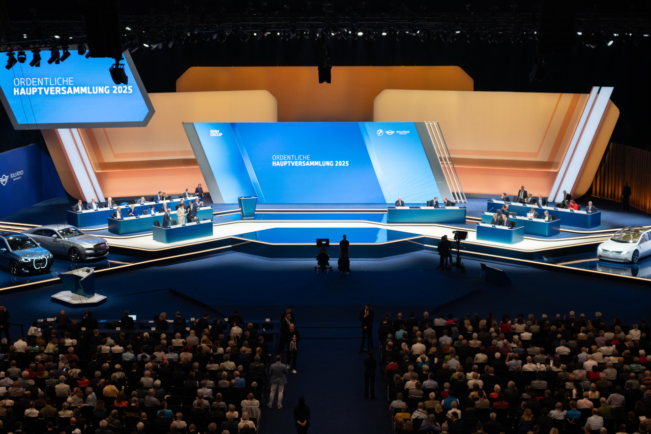 View of the stage at the 2025 BMW Annual General Meeting with podium, board members, vehicles, and audience in a large hall.