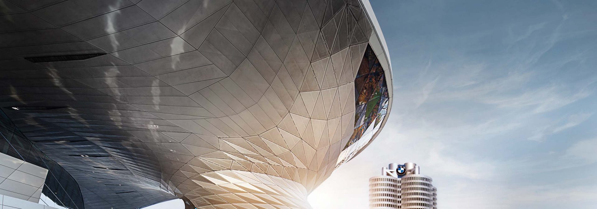 Links im Bild die BMW Welt mit futuristischer Metallfassade, rechts im Hintergrund das BMW-Vierzylinderhochhaus bei Sonne.