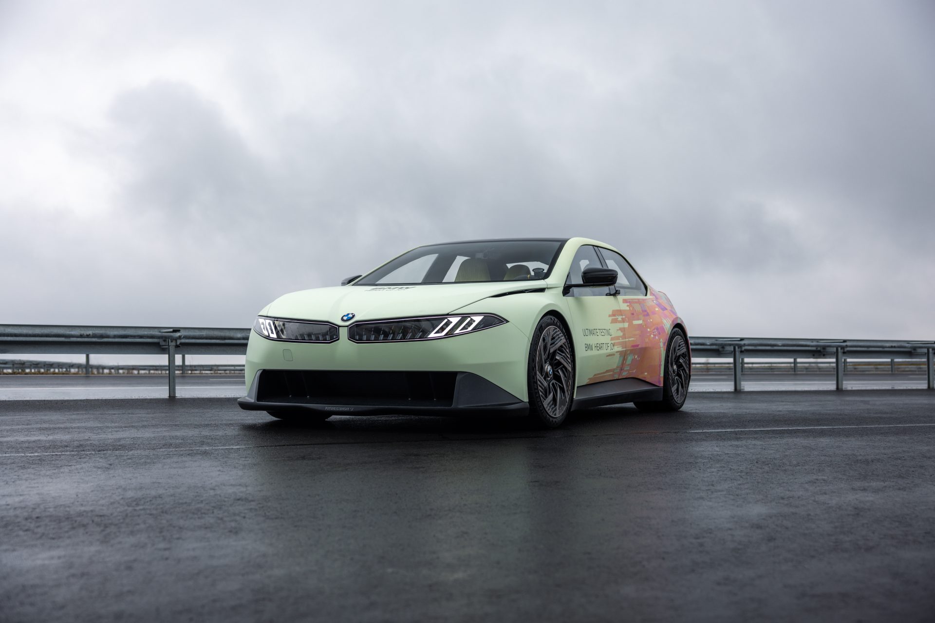 Light-colored BMW vehicle with colorful side decals parked on wet pavement under cloudy sky in front of guardrails.
