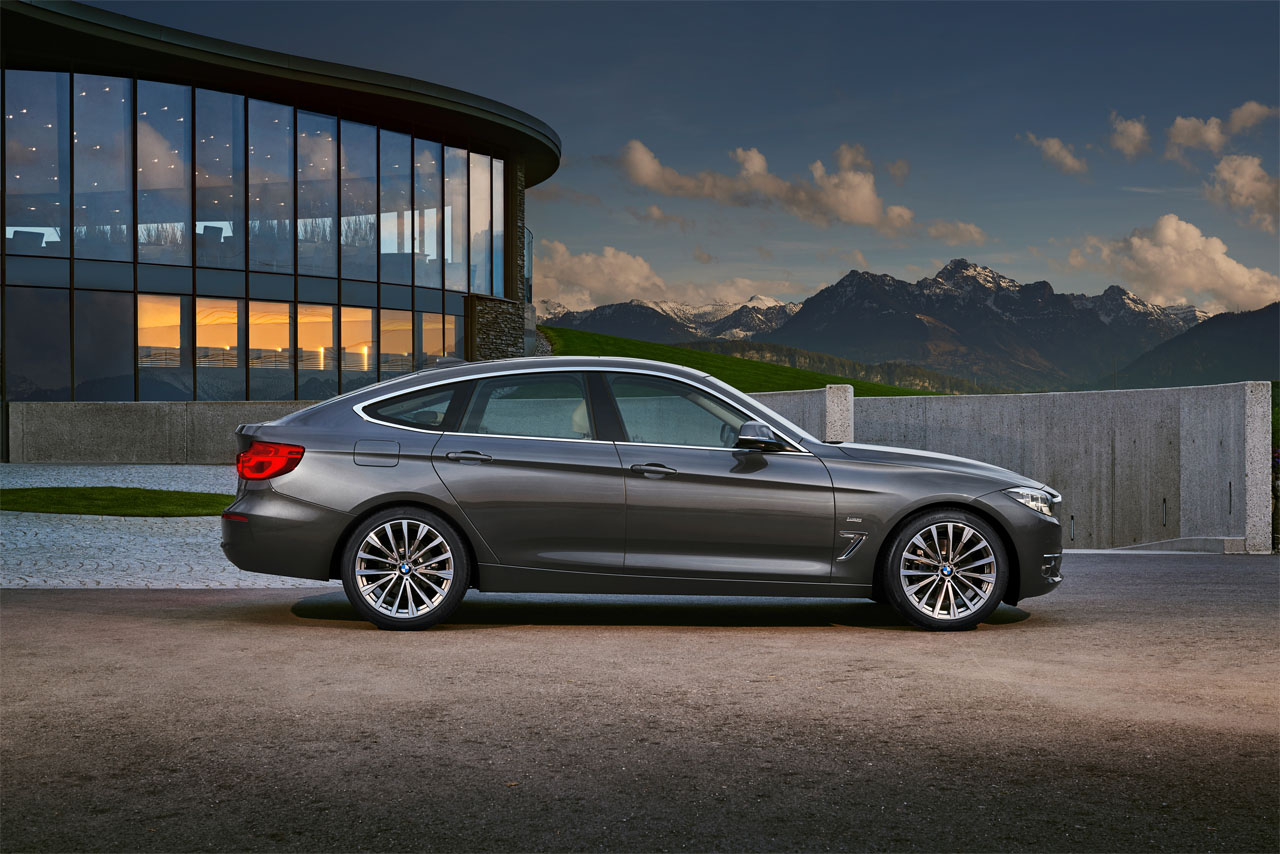 Side view of a dark grey BMW F34 Gran Turismo, seen from the right, against a backdrop of modern architecture and mountains.