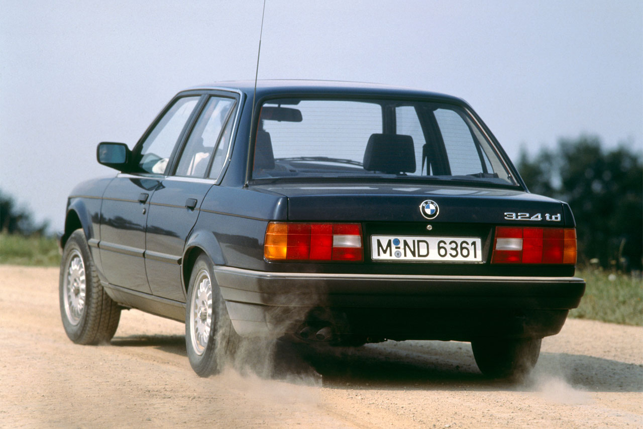 A classic diesel in motion: The rear and side of a dark BMW E30 324td as it throws up dust on an untarred track.