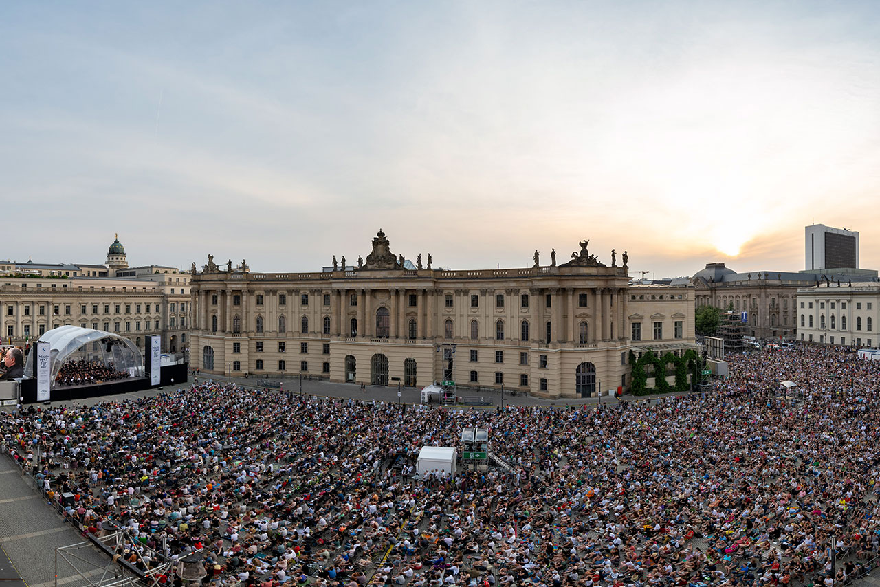 A very large crowd stands in front of a stage in the centre of a city between historic buildings.