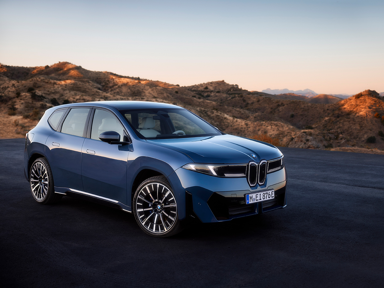 Der neue BMW iX3 in blau auf einer leeren Straße mit Sanddünen im Hintergrund.