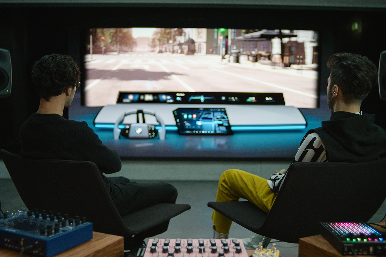 Zwei Sounddesigner der BMW Group blicken im Studio auf eine Leinwand mit virtueller Frontscheibenansicht und das BMW Panoramic iDrive, umgeben von Soundtechnik.