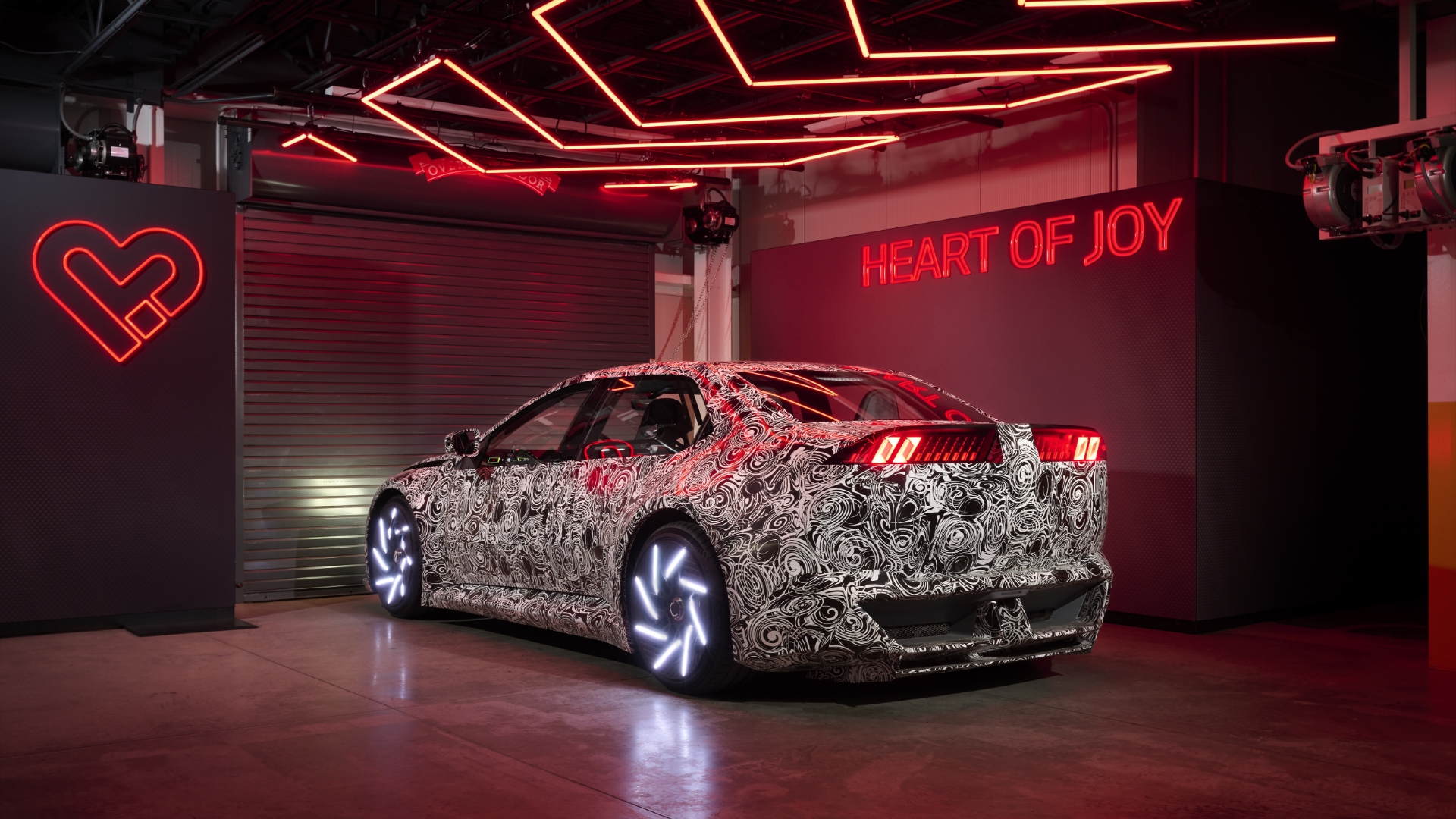 Ein BMW mit Tarnfolie steht vor einem geschlossenen Rolltor in einem stimmungsvoll beleuchteten Raum. Rechts an der Wand leuchtet in Neon der Schriftzug „Heart of Joy“, links ein Herz.