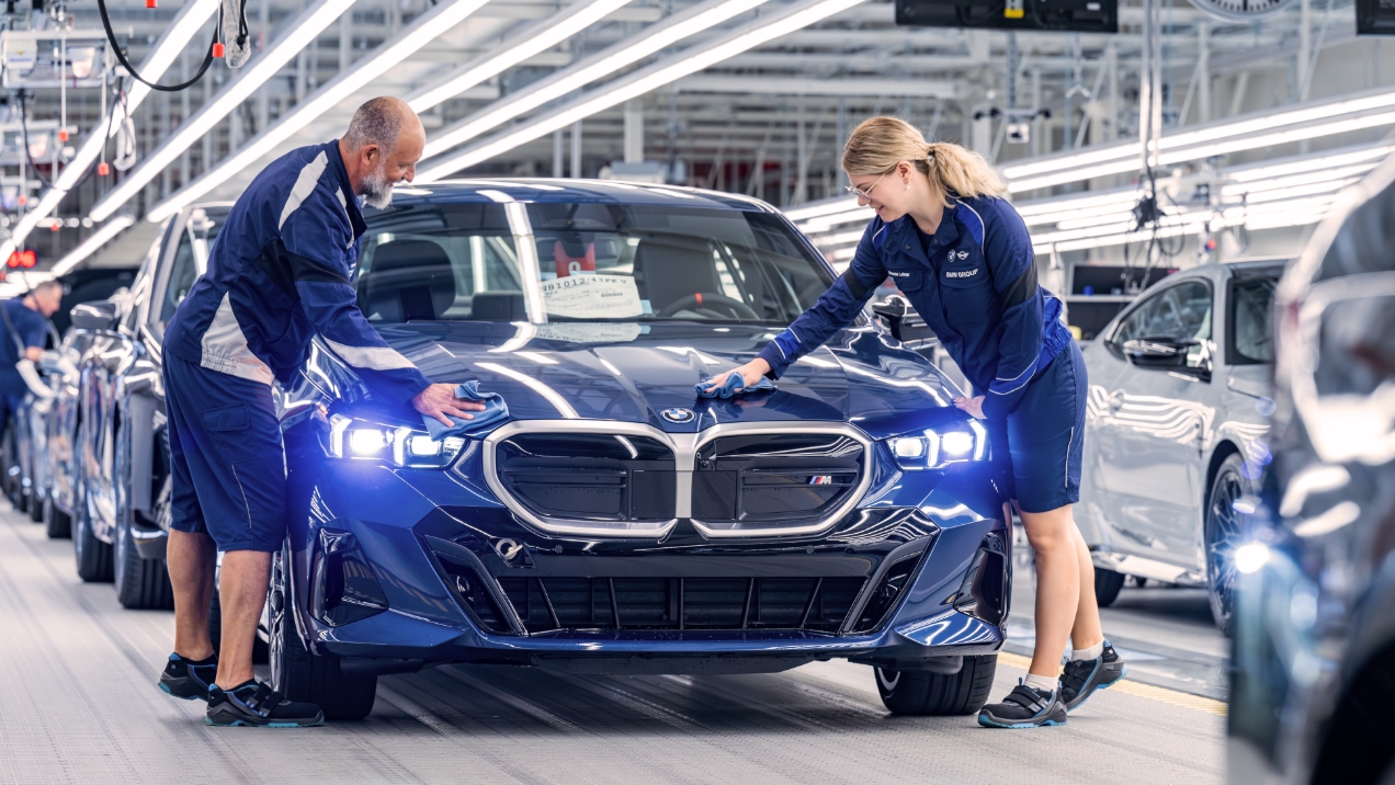 Zwei Menschen stehen links und rechts von einem dunkelblauen Auto der Marke BMW auf einer Produktionslinie und polieren die Kühlerhaube mit Tüchern.