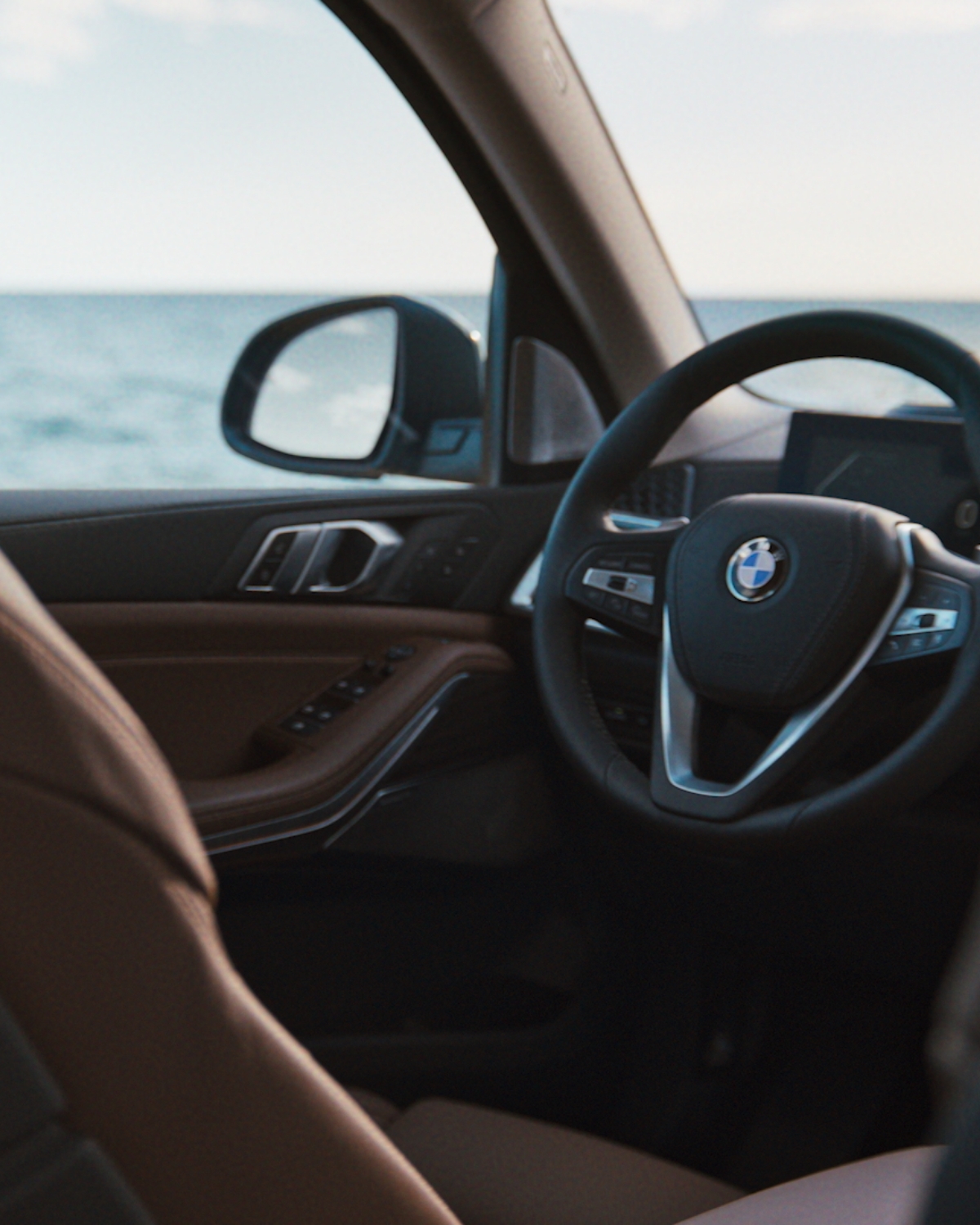 Schräge Perspektive in das Cockpit eines BMW auf das Lenkrad und den Außenspiegel mit dem Meer Hintergrund.