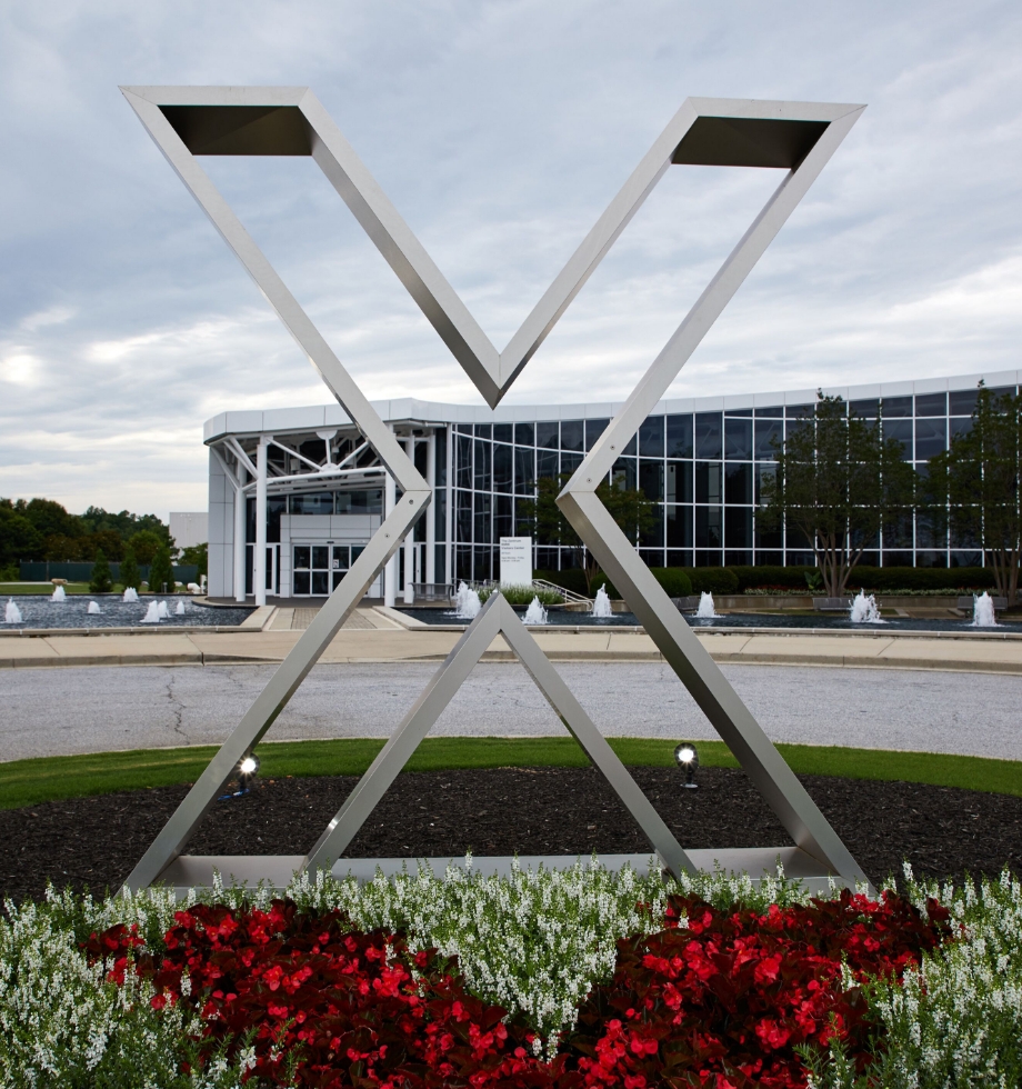 Eine Statue in Form eines großen "X", steht in einem Blumenbeet vor einem modernen Gebäude.