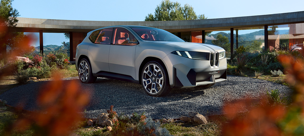 Das Visionsfahrzeug „BMW Vision X“ steht auf einem Kiesplatz, eingerahmt von dunkler Vegetation im Vordergrund.