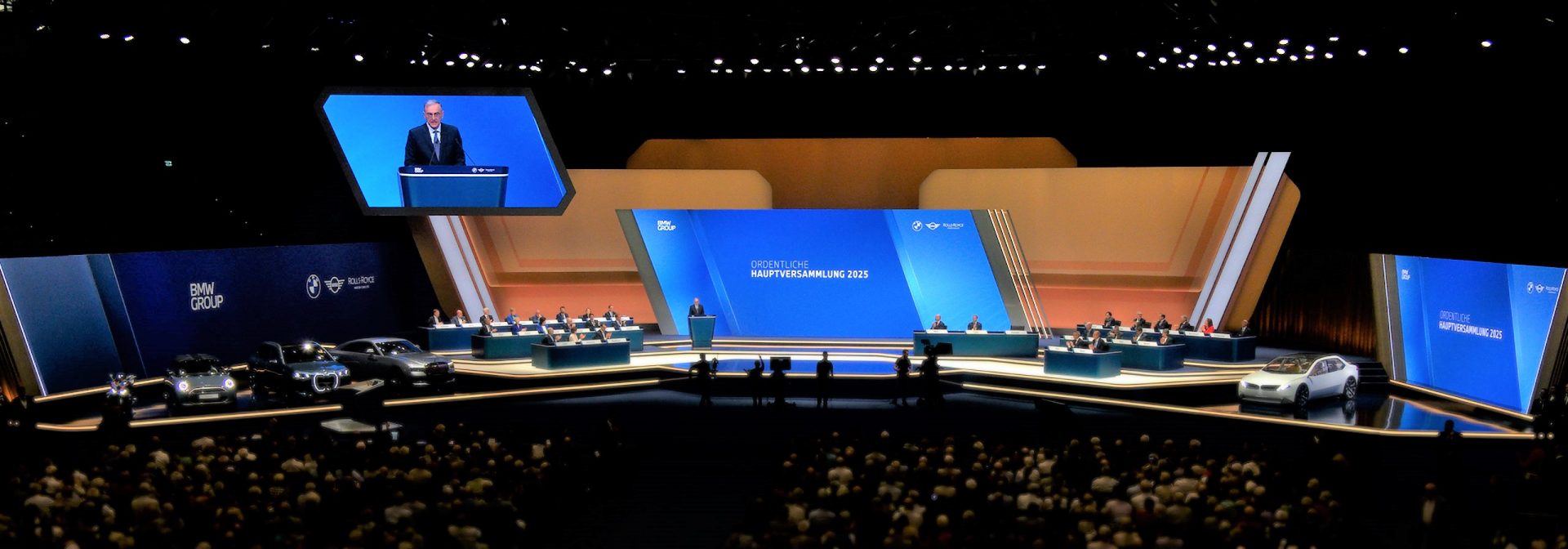 Bühne der BMW Hauptversammlung 2025 mit Rednerpult, Vorstandsgremium, Präsentationsflächen und ausgestellten Fahrzeugen.