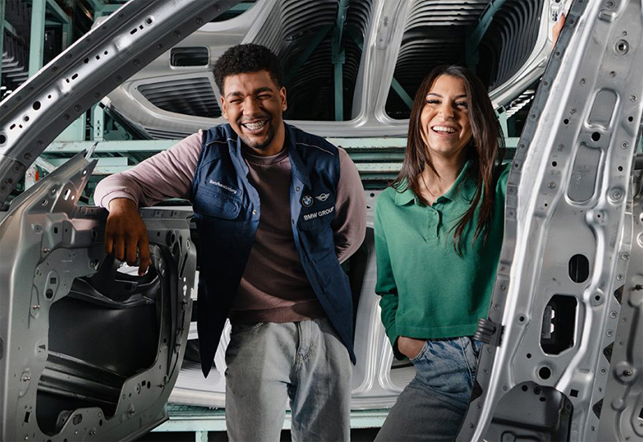 Zwei junge BMW Mitarbeiter lächeln durch eine Autokarosserie in die Kamera.