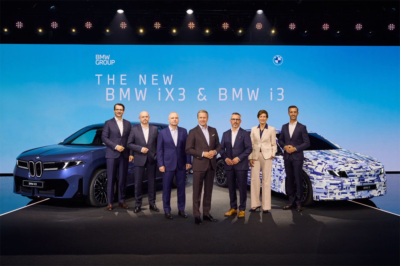 Sieben Personen in Anzügen stehen vor zwei BMW-Fahrzeugen, einem blauen BMW iX3 und einem getarnten BMW i3, vor einer blauen Wand mit der Aufschrift 'THE NEW BMW iX3 & BMW i3'.
