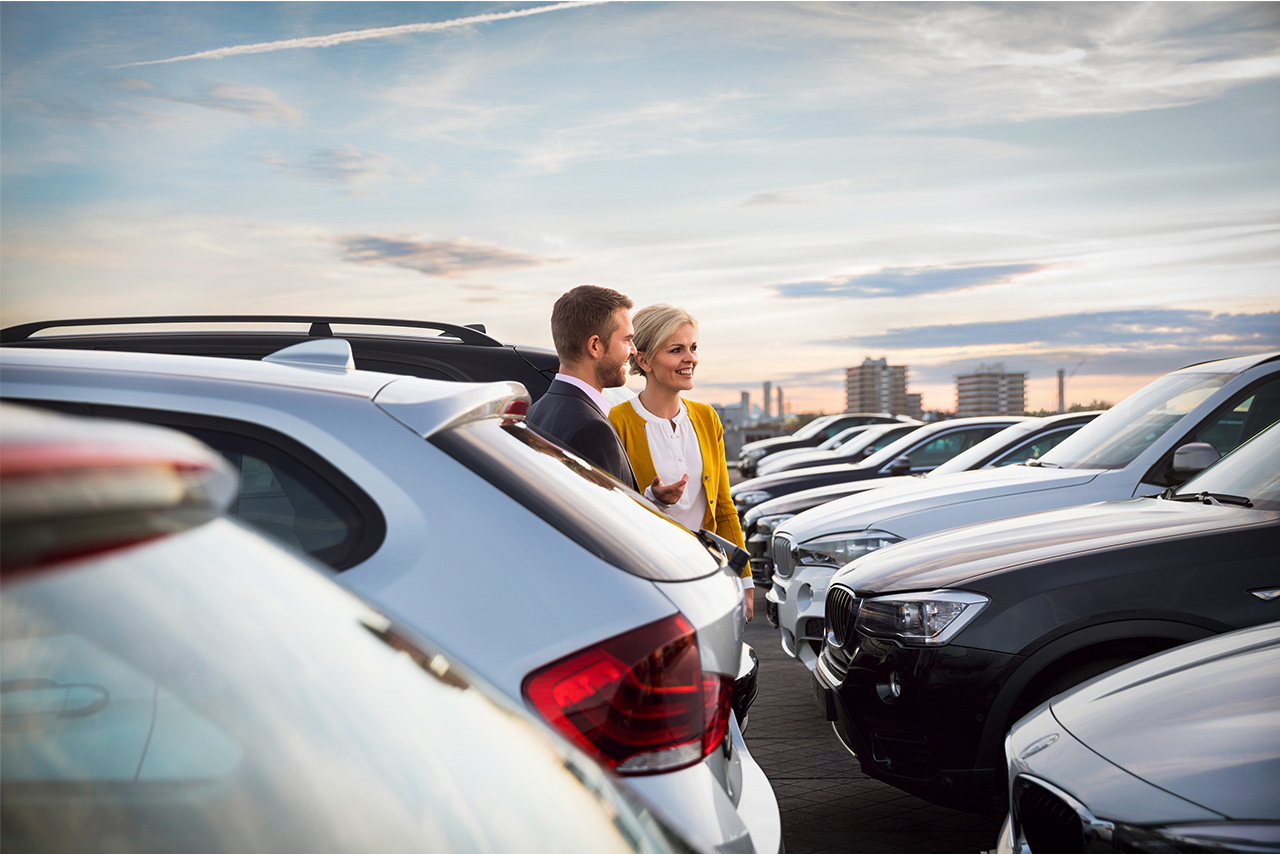 Ein Alphabet Mitarbeiter läuft mit einer Kundin über einen Parkplatz mit vielen BMW Fahrzeugen.