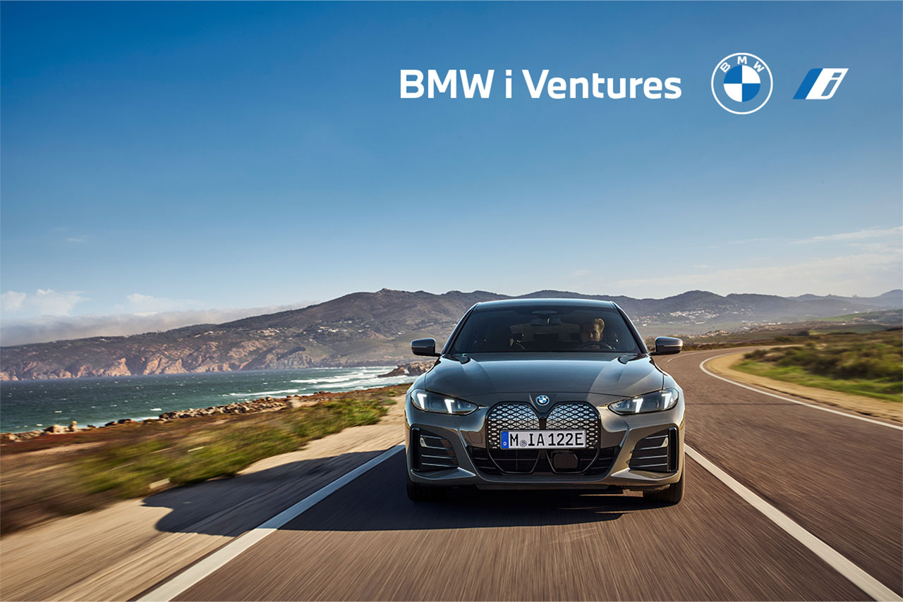 Ein grauer BMW i4 fährt auf einer kurvenreichen Küstenstraße. Im Hintergrund sind Berge und das Meer sichtbar. Text: 'BMW i Ventures' und das BMW-Logo.