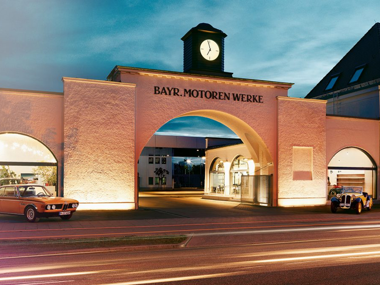 Historic factory gate with clock and the inscription ‘Bayr. Motoren Werke’ at dusk, flanked by two classic BMW vehicles.