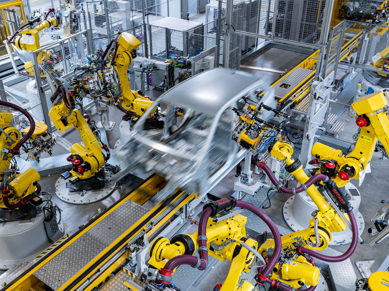 A manufacturing facility with multiple yellow industrial robots working on a silver car body moving along a conveyor belt.