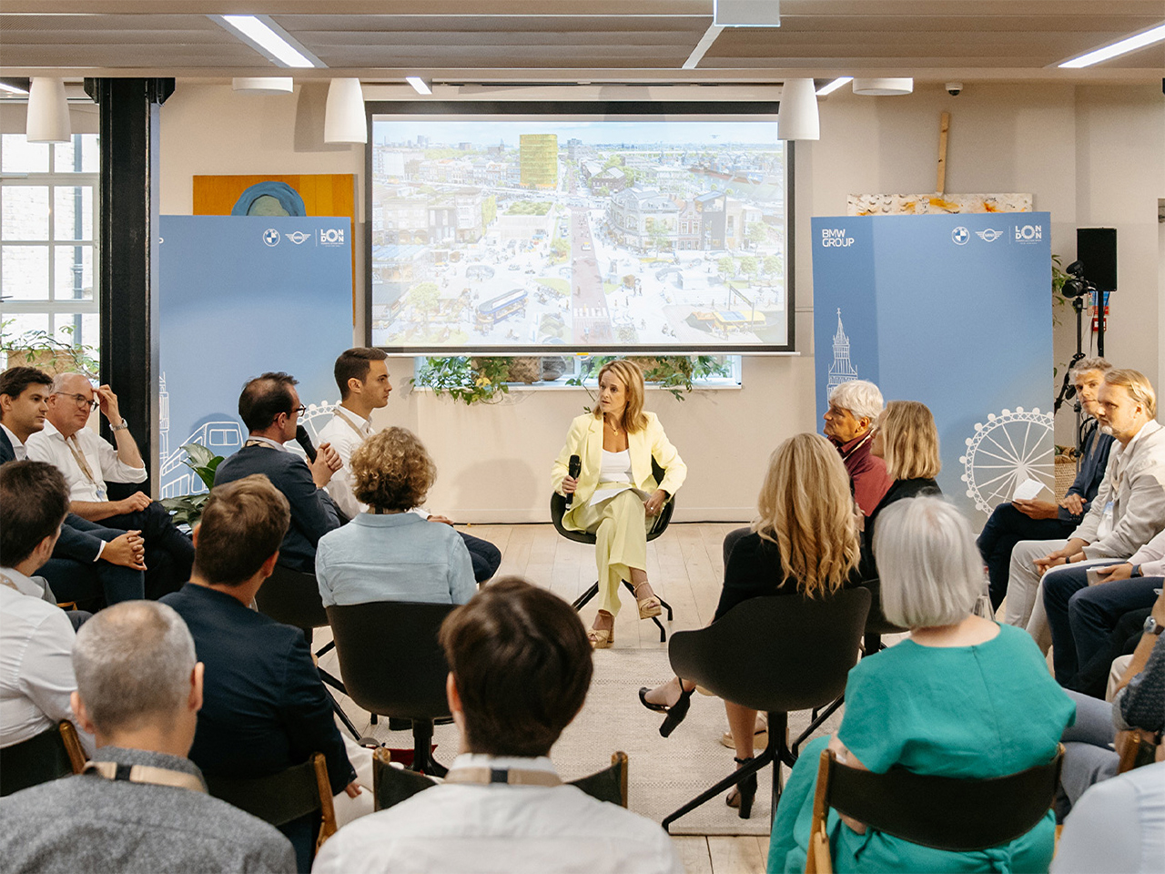 Several people sitting together, listening to a talk by a woman, behind her your can see a city projected onto a whiteboard.