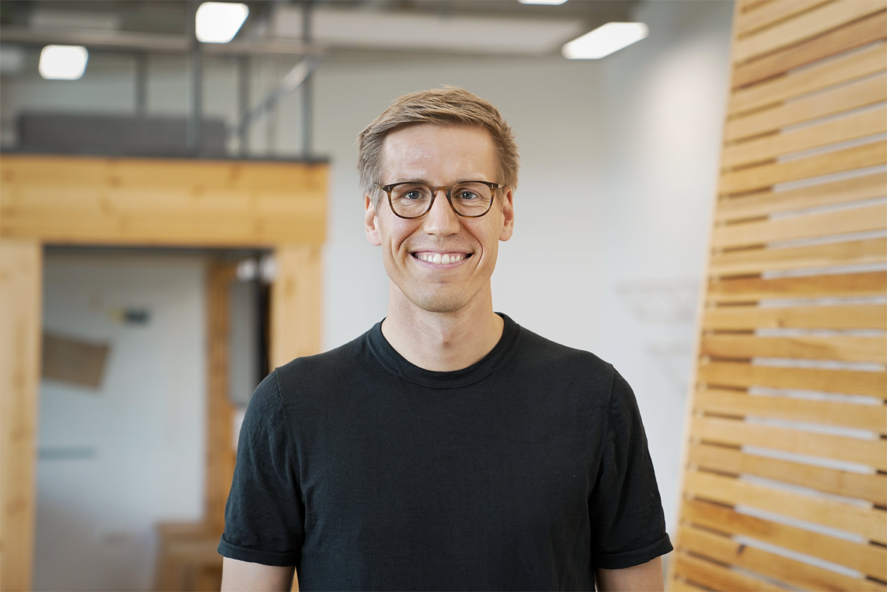 Man with short blonde hair wearing a black t-shirt stands in a room with wooden shelves and a modern, minimalist design.