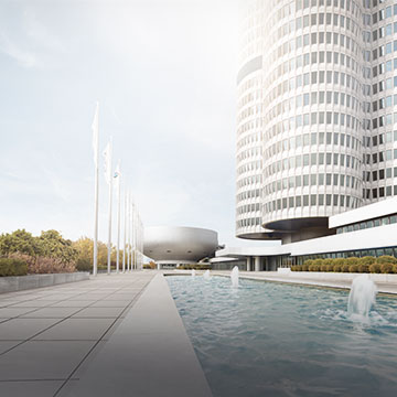 BMW Four-Cylinder Tower on the right, with flagpoles and a water basin on a paved area to the left under a bright sky.
