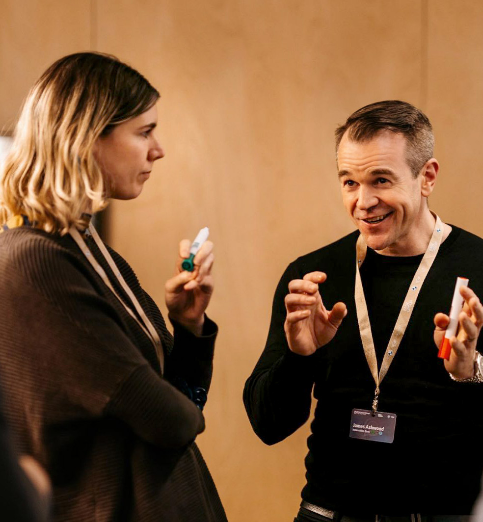 A person with a name badge is talking to another person, both holding whiteboard pens.