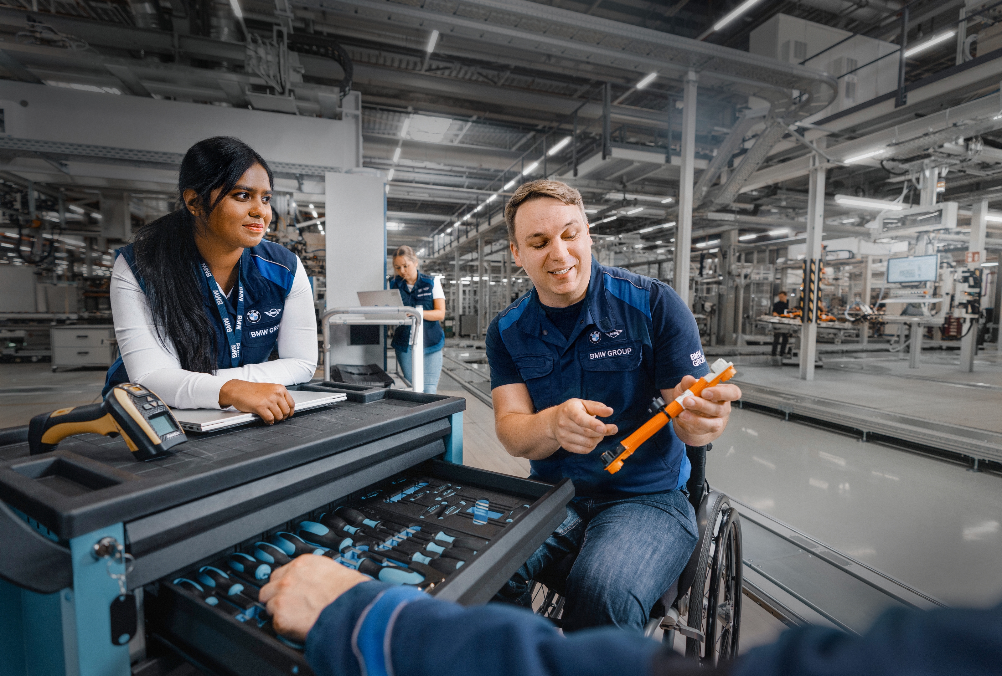 Employees of the BMW Group are standing at a workbench in a production hall. A man shows a woman a tool.